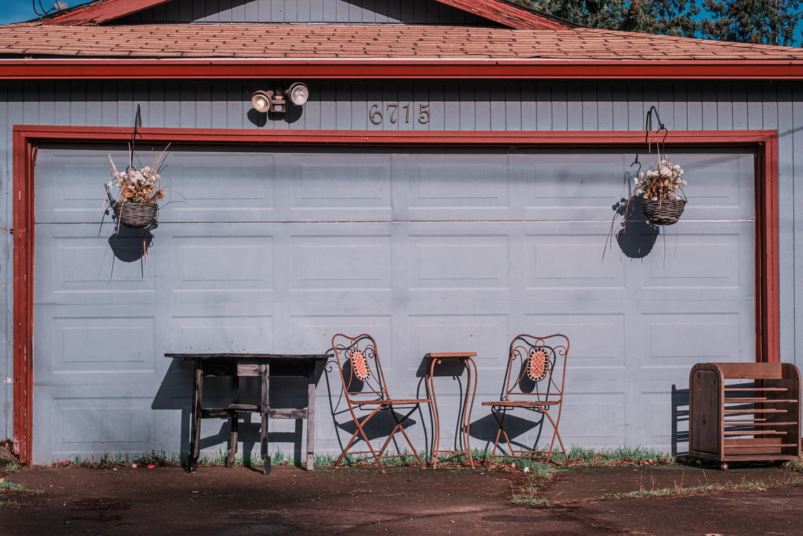 A pair of wooden and wire chairs next to a faded, chipped table in front of a faded blue and orange garage door