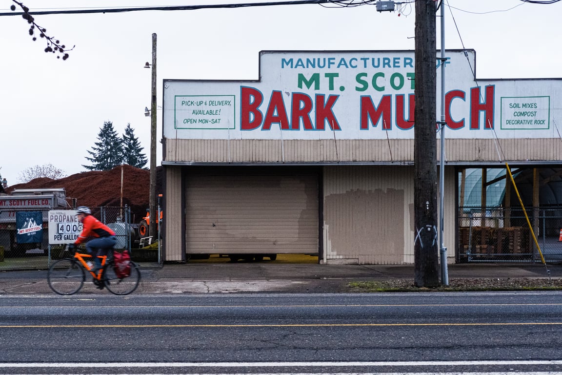 A bicyclist in an orange jacket is blurred in motion as he passes in front of a building with a sign that reads &ldquo;Mt. Scott Bark Mulch.&quot;