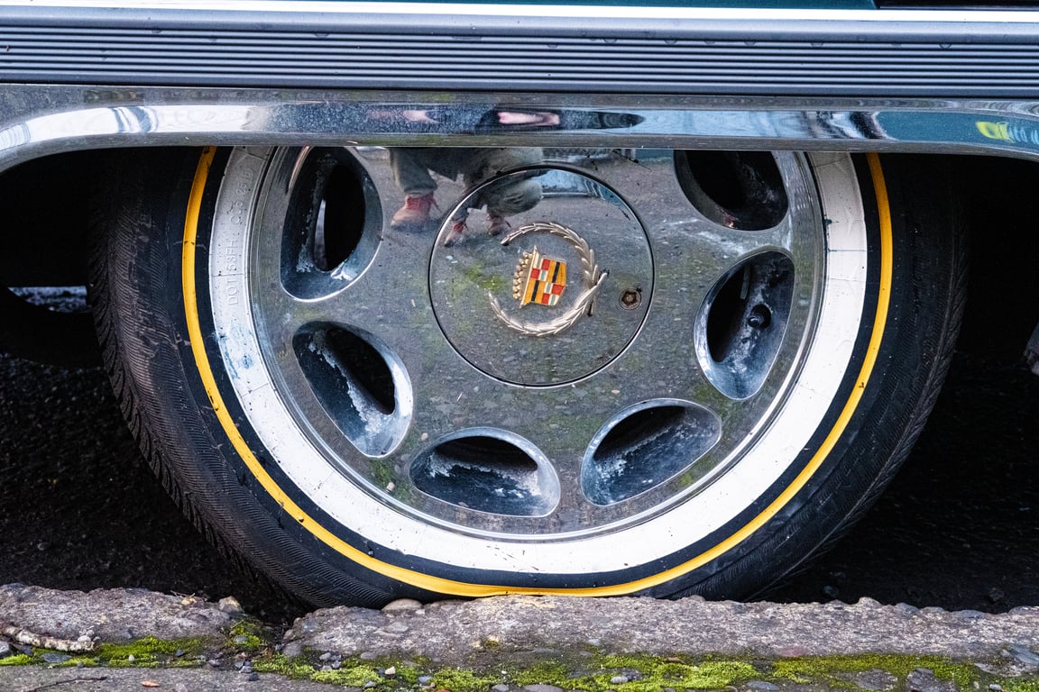 A Cadillac hubcap. The tire has a bright yellow stripe. You can see the photographer&rsquo;s feet reflected in the hubcap&rsquo;s chrome.